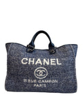 A Chanel tote bag in a blue and gray herringbone tweed fabric, featuring white embroidered 'CHANEL' text and logo on the front, dark blue leather handles, and silver-tone hardware.