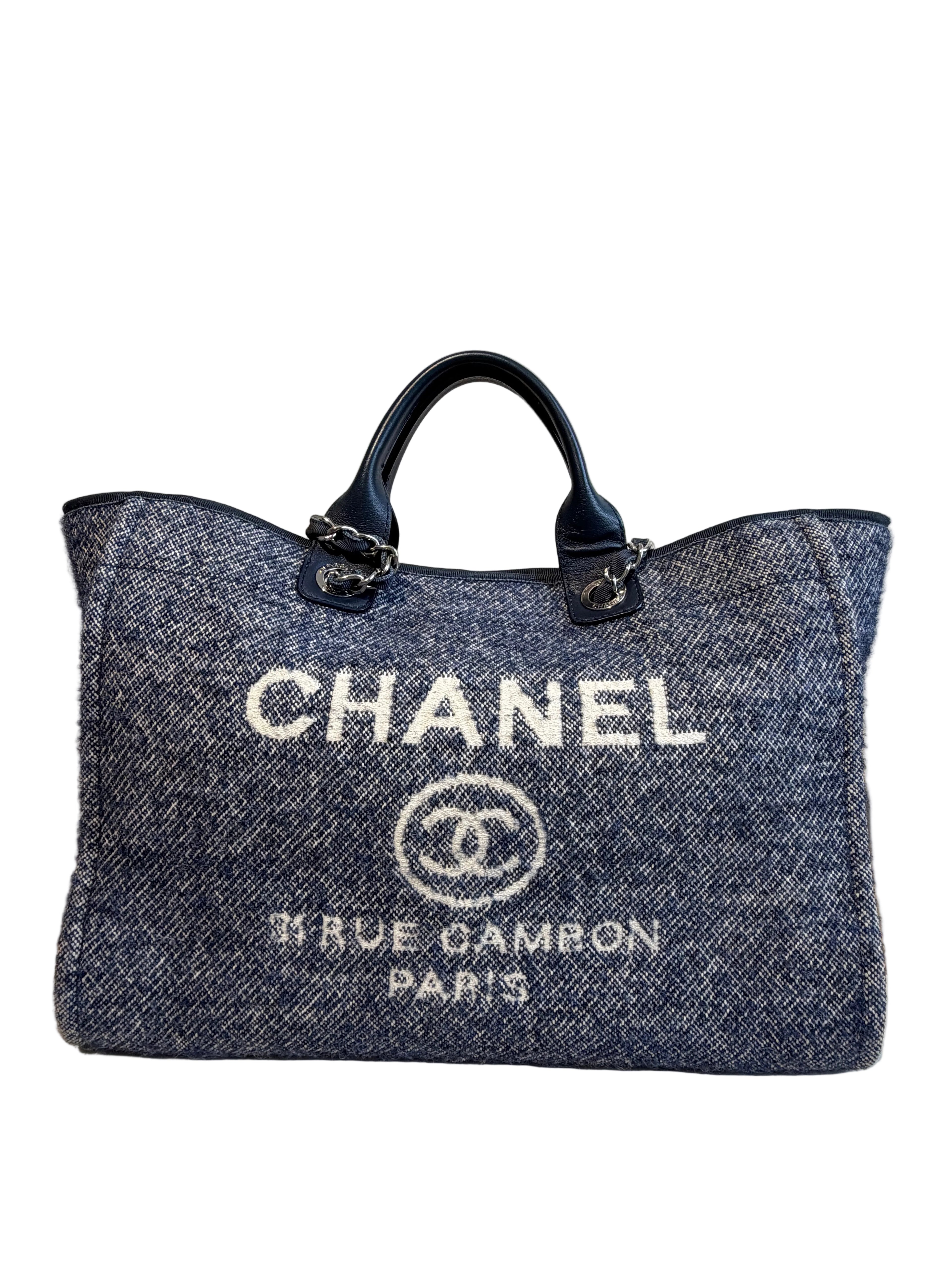 A Chanel tote bag in a blue and gray herringbone tweed fabric, featuring white embroidered 'CHANEL' text and logo on the front, dark blue leather handles, and silver-tone hardware.